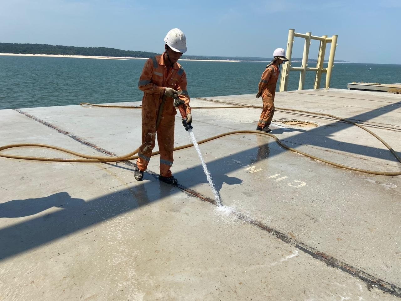 Trabalhadores com equipamentos de proteção lavando superfícies no convés de uma embarcação.